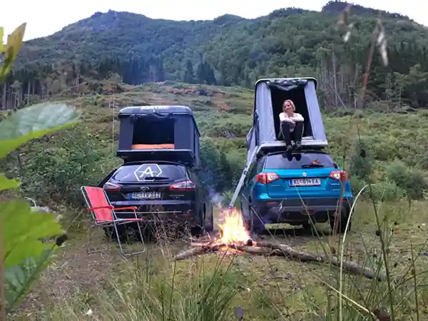 Zwei Autos mit Hartschalendachzelten. Frau sitzt im Dachzelt Feuer Natur