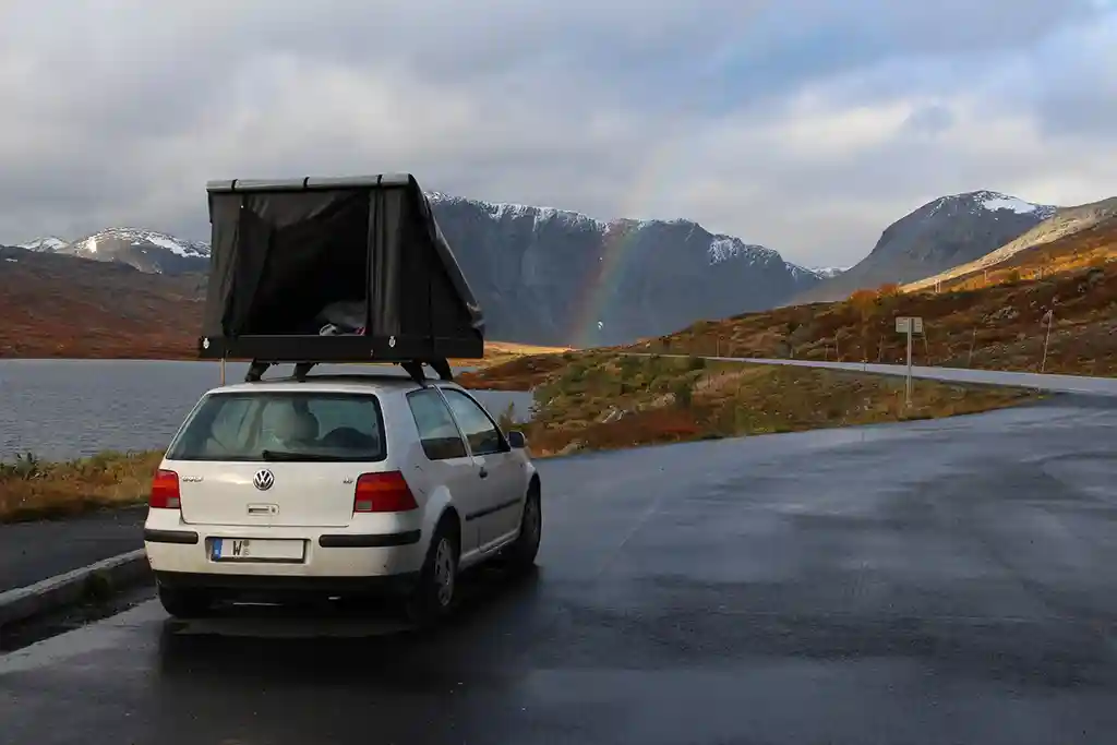 regenbogen weisses auto dachzelt wolken blauer himmel strasse baeume huegel