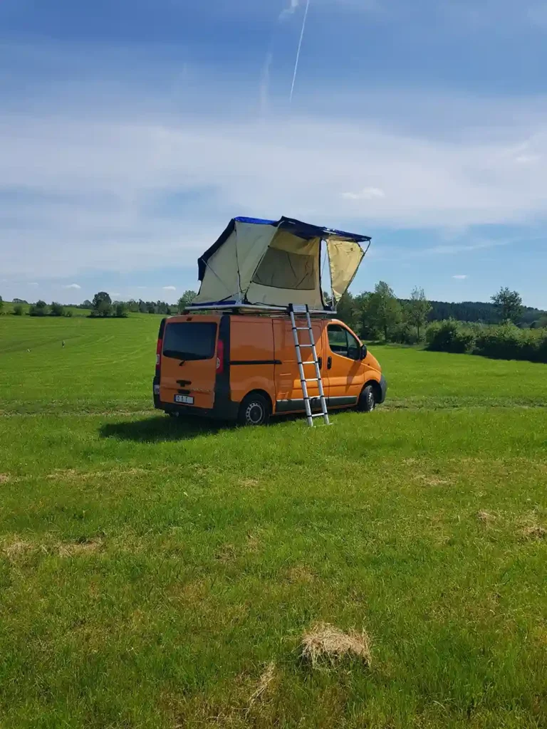 selbst gebautes dachzelt auf orangem auto montiert wiese blauer himmel